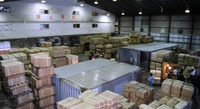 Red letters are written on the high upper wall, towards the ceiling of a warehouse, saying "No Smoking," in a scene right before a firey explosion in the factory. The warehouse is filled with smaller boxes and few large industrial storage containers. a worker drives a forklift shifting boxes.