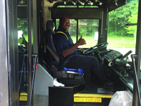 Color photograph taken through open door of bus, showing driver, an African American man, smiling and giving “thumbs up” sign.