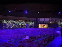 Exhibit photo: Dark room with a purple illuminated floor. The floor is in the foreground and there are small sculptures of buildings in the front of wall projections.  On the walls is the projection "The Secret Garden" by artist Stephanie Dinkins. The projection shows two Black women, one on the left and one on the right. Behind them are cotton, okra, and green leaves. Above the projection is a black sky and a few purple lights.