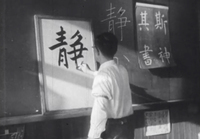 A teacher in a classroom draws black calligraphy on a whiteboard set against the chalkboard with white calligraphy written on it with paper attached to it with white calligraphy, in black and white cinematography.
