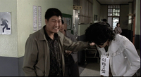 Two individuals converse in a hallway, with s sign worn around one of their necks with black calligraphy printed on it. Signs with black and red calligraphy printed on them are posted on the wall.