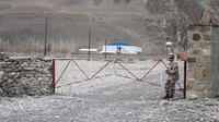 Two young Afghan men stand on a dirt road in Shughnan, Afghanistan, which is on a small hill that overlooks the Panj River. The view shows the Panj River, the Tem border-crossing area on the outskirts of Khorog, the road across from the river, and small farmlands and adobe houses.