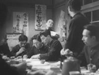 A classroom of young men sitting around a table in front of black calligraphy on various banners on the wall, in black and white cinematography.