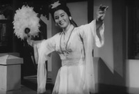 A black-and-white film still in a medium shot. It shows a female figure in a traditional-style robe dancing with her arms stretched out, holding a fluffy feather fan in her   hand.