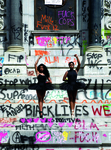Two young female Black dancers, one with long hair and one with short hair or hair pulled back, appear in front of a monument covered in colorful layered graffiti which reads in part “save black lives,” “Stop white supremacy,” “Fuck pigs,” “FTP,” and “BLM.” The dancers are dressed in black leotards with classical tutus and are posed en pointe in pointe shoes. Each one has a single arm lifted overhead. Their extended arms are in a ballet position with their hands clenched in fists.