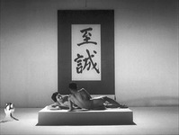 A film still of a large calligraphy scroll hanging behind a naked man and woman, laying together on a raised platform in a sparse room.