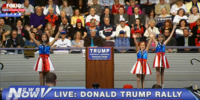 Three young girls in shiny red, white, and blue dresses raise their arms in unison. The girls appear on either side of a podium with a sign that reads “Trump” and “Pensacola, Florida.” An audience sits behind them in the stands.