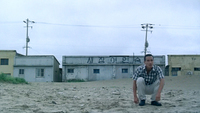 A man crouches on the beach. On the building behind him is painted a sign. The hangul, "새장여인숙" (Saejang Inn) is a play on words. Inserting space ("새장 여인 숙") converts the sign to something like "Woman in a Cage." Ironically, this indicates the position of the female character with him.