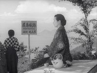 A woman sits on a semi-sunny ledge next to a teapot and three cups, and with her are two people standing and overlooking a large body of water with mountains and trees in the far background. There is a sign for an inn between the seated woman and the standing people.