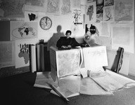 A person sits at a desk overflowing with maps. A variety of other maps and papers cover the walls and floor.