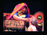 On stage, the character Ibedon paints Dule Wagwa’s face as Dule Wagwa kneels.