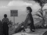 A woman sits to tea in the outdoors in black and white cinematography. In front of a high mountainscape view, with a sign with black calligraphy in front of her.