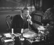 A man sitting at a desk, talking on the telephone while reading a newspaper.