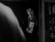 Dim, black and white image of a woman standing at a doorway holding a white piece of paper with calligraphy on it. To the right of the doorway, the wall has a large banner of calligraphy.