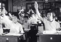 A classroom of students raise their hands in front of a banner with white calligraphy, in black and white cinematography.