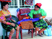 Sisters Lois and Domatila De León Kantule sitting down while wearing their brightly colored work, with more artwork hung on a chair between them.