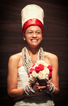 A performance still depicting a mixed-race woman wearing a tall white “African” hat with beads around her neck and wrists, carrying a red-and-white bouquet and smiling awkwardly.