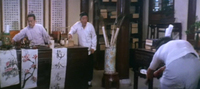 A scene set in a study. All the implements of calligraphy are on the desk, with three strips of paper drying on its side. One of the strips has wispy calligraphy. Behind the desk and teacher are three scrolls dense with calligraphic writing. Other scrolls are stored in a jar next to the desk.