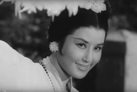 A black-and-white film still in a close-up shot. It features the same female figure disclosing her beautifully made-up face from behind the fluffy feather fan at the ending pose of her dance.
