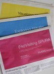 Photo of three exhibition newspapers—one pink, one yellow, one blue-fanned out on a table, and one from each year of the exhibitions.