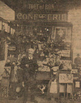 Photograph of Georges Méliès standing in front of a small kiosk overflowing with toys. Kiosk sign reads, Tout est Bon Confiserie.