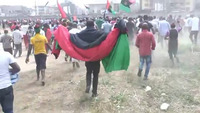 Biafra protesters in southeast Nigeria, 2016. Demonstrators walk in a wide formation over grass toward a point away from the camera. Other protestors are waving Biafran flags. In the foreground of the picture, a protester walks with a Biafran flag draped across their shoulders.