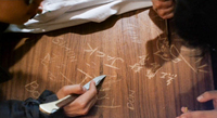 A birds-eye close-up of the hands of two young grifters scratching calligraphy characters, the English names "Jack" and "Simon," and a pictograph onto a table with switchblades.
