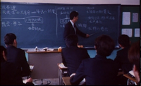 A teacher instructs a classroom of students in front of a chalkboard with white calligraphy written on it.