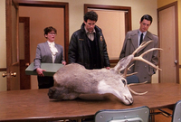 A woman and two men stand in a wood-­paneled office room, looking at an overturned taxidermy deer head on a table in the foreground.