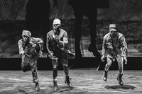 Fig. 18. Three male performers playing African soldiers in World War I. They march, desperate, exhausted, determined, shadows looming behind them on a screen. Black and white photo.