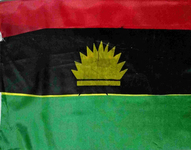 The flag of Biafra. The flag has three horizonal panels in red, black, and green running from top to bottom. In the center of the flag, within the black panel, there is an icon of a golden rising sun on a golden bar.