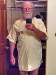 A gray-­haired man with bushy beard, is taking a selfie with his phone while looking into the bathroom mirror. He wears a knee-­length white smock numbered 5486 on the chest.