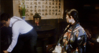 A man and woman talk in a dark wood room, with a calligraphy scroll on the wall behind them.