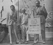 Paul Whiteman on an outside stage, flanked by Carl Laemmle Jr. and Carl Laemmle, speaking at a microphone, behind a sign: “Let’s Tell the World about Paul Whiteman and the ‘King of Jazz