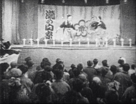 A crowd watches a stage performance. Calligraphy is visible on the banner behind the performers.