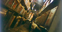 The image is canted to the right. A storehouse is filled with large pots with red calligraphy written on them on three stories of shelving on both sides of a narrow hallway. The hallway ends in two tall glass windows. A man is on a ladder on the right side in the midground, observing one of the pots.