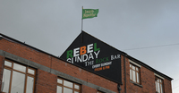 A brick building bears a broadside event advertisement. A flagpole on the building’s roof flies a green 1916 Irish Republic flag.