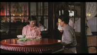 Image of two people sitting at a round wooden table in a restaurant, doing prep work with a bowl of greens. One of the people is folding a piece of paper as the other watches. Through a window, in the deep background, there are banners on the wall in pink and red with black calligraphy written on them.