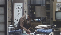 A man works over a pile of paper and fabric in his home. Calligraphy is visible on signs and posters in the background.