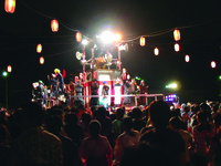 Fig. 10. A photograph of a nighttime performance. Several people perform a choreographed dance routine on a raised platform, surrounded by a crowd. Red lanterns are hung leading from the platform, over the crowd.