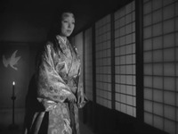 A black-and-white film still, showing a female figure dressed in a shiny silk kimono, emerging from the darkness and standing in front of a Japanese-style side door. Behind her is a folding screen painted with a crane and a candle stand holding a burning candle below.