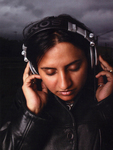 A brown-skinned woman touches her hands to her large headphones, looking down and listening contemplatively.