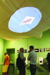 A white parachute hangs from the ceiling. Projected onto the parachute is an image of a white kite with a person’s face flying in a blue sky.