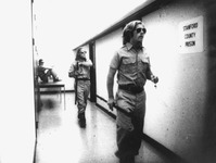 Three mock-­guards in the Stanford prison corridor, all wearing dark shades. The first, in the foreground, walks toward a cell, holding a whistle and a cigarette. Behind him, a second man holding a long baton. A third man sits on a table behind them, his back against the wall.