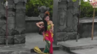 Men, seated, chant as an elegantly dressed woman enters from a gate at the side.