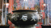 A large oval bronze incense holder, with silver calligraphy embossed on it, fills the frame. Many incense emerge from the top of it. At the bottom left and right, the corner of two rectangular incense holders flank it, with many incense emerging from inside it. A light, diffuse incense smoke lingers generally around the tops of the incense.