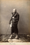 A sepia photograph of a blind masseur dressed in a robe and wearing raised wooden clogs. He holds a flute-like whistle to his mouth while the other hand holds a walking staff. The caption reads “Blind Rubber.”