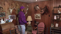 Man wearing a purple athletic jacket stands in a wood-­paneled room filled with chairs, framed photographs, and taxidermic mounts. His side is turned to the camera, and he faces a large boar’s head mounted on the wall. His hand is reaching out to touch a deer mount next to the boar.
