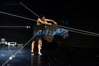 A dancer hangs black pieces of clothing on wires intersecting in the middle of a black box proscenium stage. A number in the billions representing the debt of Greece is projected in blue on the fabric.