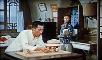 In a residential setting, a man at a table builds a wooden model of a milling machine. A woman looks on, smiling.